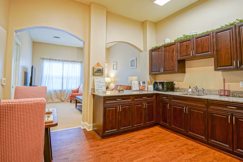 Interior view of a senior living facility showing a kitchen area with dark wooden cabinets, granite countertops, a microwave, and a sink. Adjacent to the kitchen is a living room area with red checkered chairs, a coffee table, and a window with sheer curtains allowing natural light to fill the space.