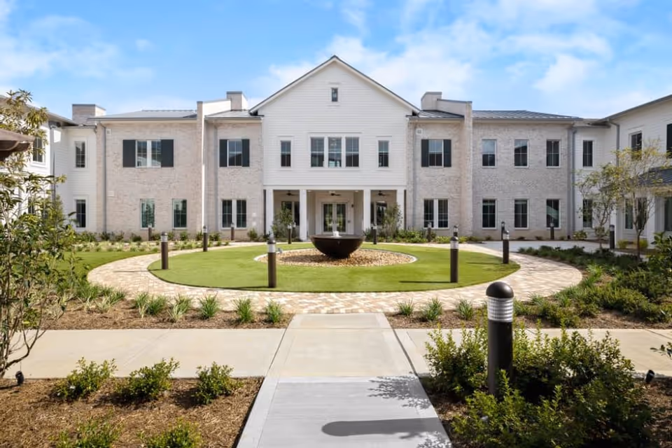 Front exterior view of The Claiborne at Gulfport Highlands building with a circular walkway and landscaped garden featuring a central water fountain under a blue sky.