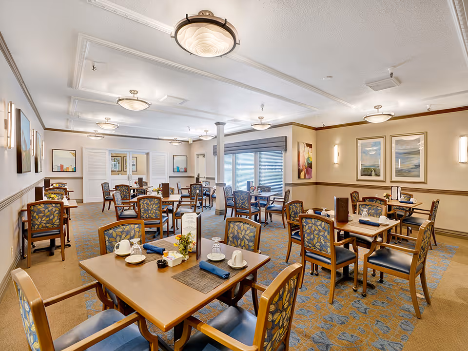 A well-lit dining room with multiple wooden tables and chairs arranged neatly. Each table is set with cups, glasses, napkins, and small flower arrangements. The room features patterned carpet, framed artwork on the walls, and ceiling lights. Large windows with blinds allow natural light to enter the space.