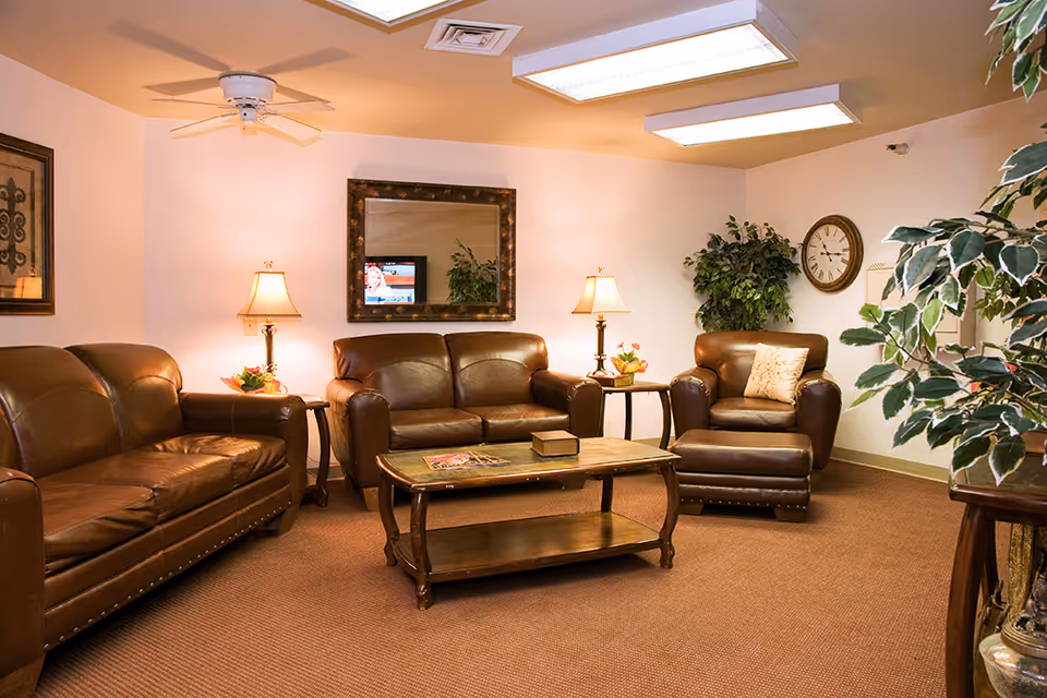 A cozy living room area with brown leather sofas and an armchair arranged around a wooden coffee table. The room is warmly lit by two table lamps on side tables and overhead fluorescent lights. There is a large mirror on the wall reflecting a TV, a clock on the wall, framed artwork, and several green plants adding a touch of nature to the space.