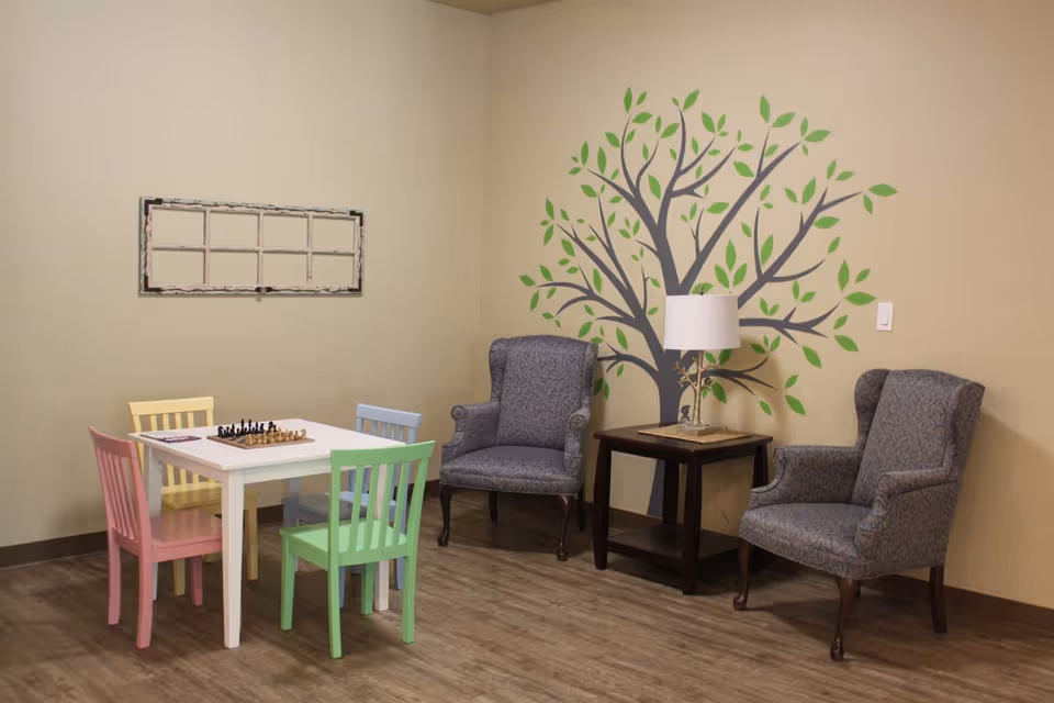 A cozy room with a small white table surrounded by four colorful chairs in pink, yellow, blue, and green. A chessboard is set up on the table. Two upholstered armchairs are placed near a dark wooden side table with a lamp. The wall behind features a decorative tree mural with green leaves.