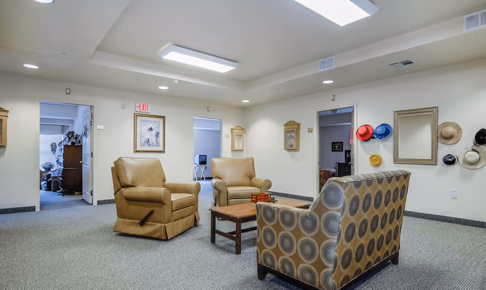 A cozy sitting area in a retirement facility with two tan armchairs and a patterned sofa arranged around a wooden coffee table. The room has light-colored walls, carpeted floor, and ceiling lights. Several colorful hats are hanging on the wall next to a large mirror. Two open doorways lead to other rooms, one with a visible desk and chair.