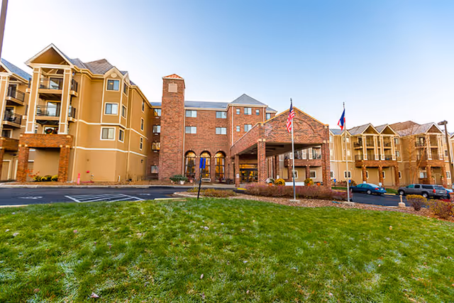 Exterior view of a multi-story senior living facility with a brick and beige facade, a covered entrance with two flagpoles displaying the American and Texas flags, a green lawn in the foreground, and several parked cars.