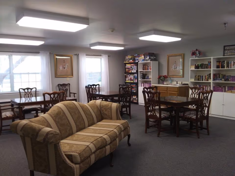 Community common room with a striped sofa, several dining tables and chairs, and bookshelves filled with books and board games.