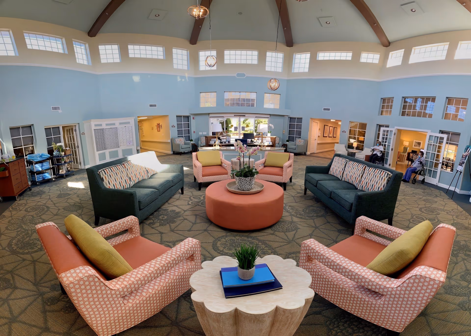 A spacious senior living facility common area with high ceilings and large windows near the ceiling allowing natural light. The room has comfortable seating including two green sofas with patterned cushions, four pink armchairs with yellow cushions, and a round pink ottoman in the center with a plant on top. There is a small wooden table with books and a plant in the foreground. The walls are painted light blue and there are multiple doorways and windows along the walls. Two elderly individuals are seated near the right side of the room.
