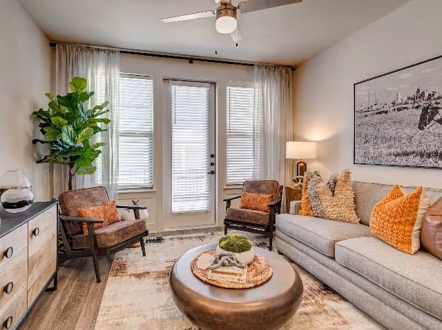 A cozy living room with a gray sofa adorned with orange and beige pillows, two brown armchairs with orange cushions, a round coffee table with decorative items, a wooden sideboard with a glass vase, a tall green plant, a ceiling fan with light, and large windows with sheer white curtains letting in natural light.