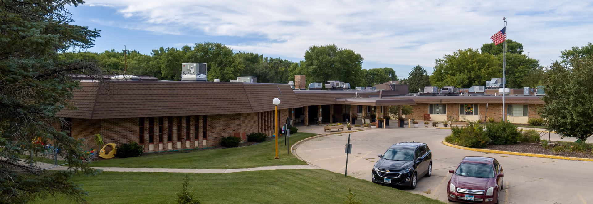 Exterior view of Morris Health Services DBA West Wind Village, a single-story brick building with a brown roof. The building is surrounded by green trees and a well-maintained lawn. There is a circular driveway with two parked cars and an American flag on a flagpole near the entrance.