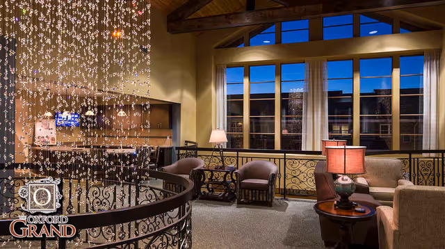 A cozy and warmly lit interior lounge area with comfortable armchairs and sofas arranged around small tables with lamps. Large windows with curtains reveal a nighttime view outside. Decorative hanging crystal strands and wrought iron railings add elegance to the space.