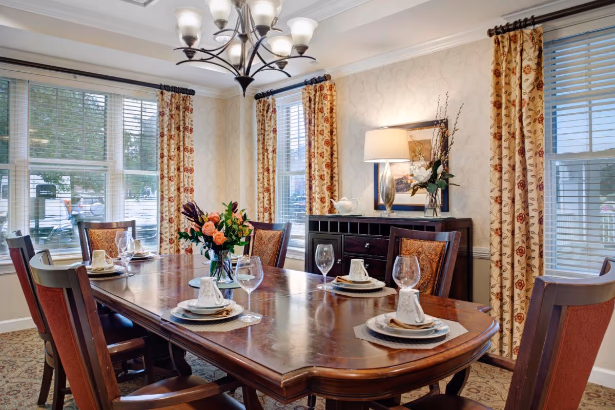 A formal dining room with a large wooden table set with plates, glasses, and a floral centerpiece, surrounded by upholstered chairs and windows with patterned curtains.