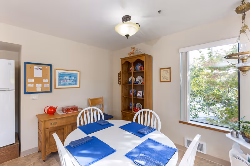 Bright dining area with a round table set with blue placemats, white chairs, a wooden cabinet, and a large window overlooking trees.