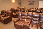 Interior view of a room with multiple brown leather reclining chairs arranged in rows, featuring cup holders in the armrests. The walls are beige with framed pictures hanging, and the floor is carpeted.
