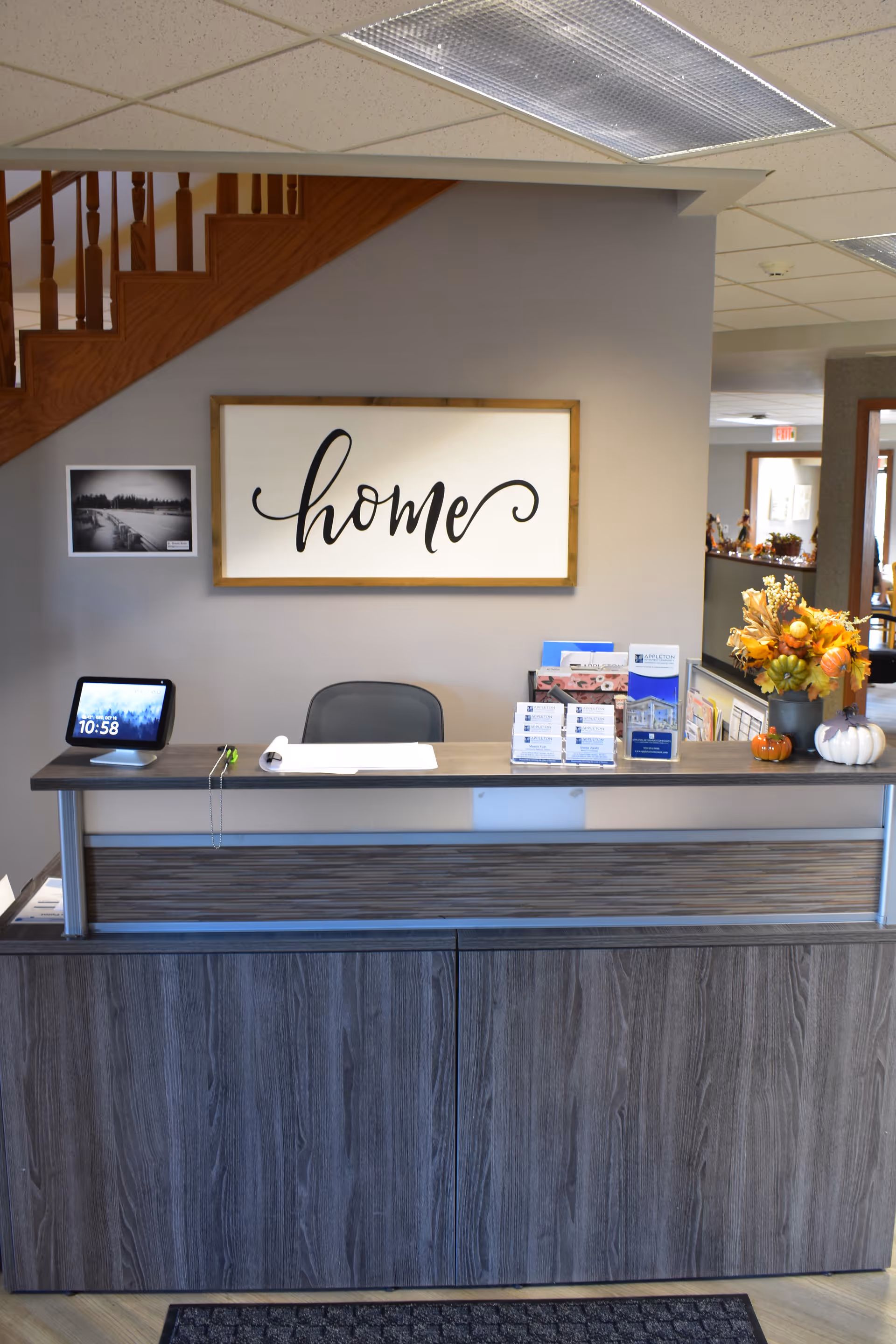 Reception desk area with a wooden front and a gray countertop. Behind the desk is a chair, a digital clock displaying 10:58, brochures, and a vase with autumn-themed flowers and small decorative pumpkins. On the wall behind the desk is a framed sign with the word 'home' in cursive script. A staircase with wooden railings is visible to the left.