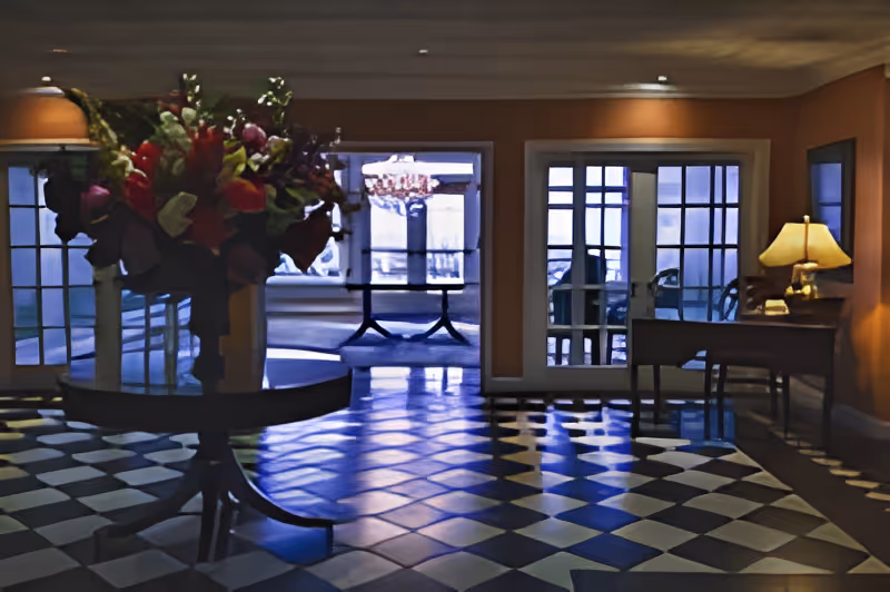 Interior view of a room with checkered black and white tile flooring, a round table with a large floral arrangement in the center, and a wooden desk with a lamp on the right side. The room has multiple glass doors and windows allowing natural light to enter.