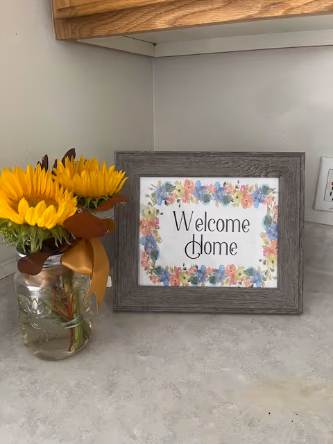 Framed "Welcome Home" sign and a mason jar of sunflowers on a kitchen countertop beneath a wooden cabinet.