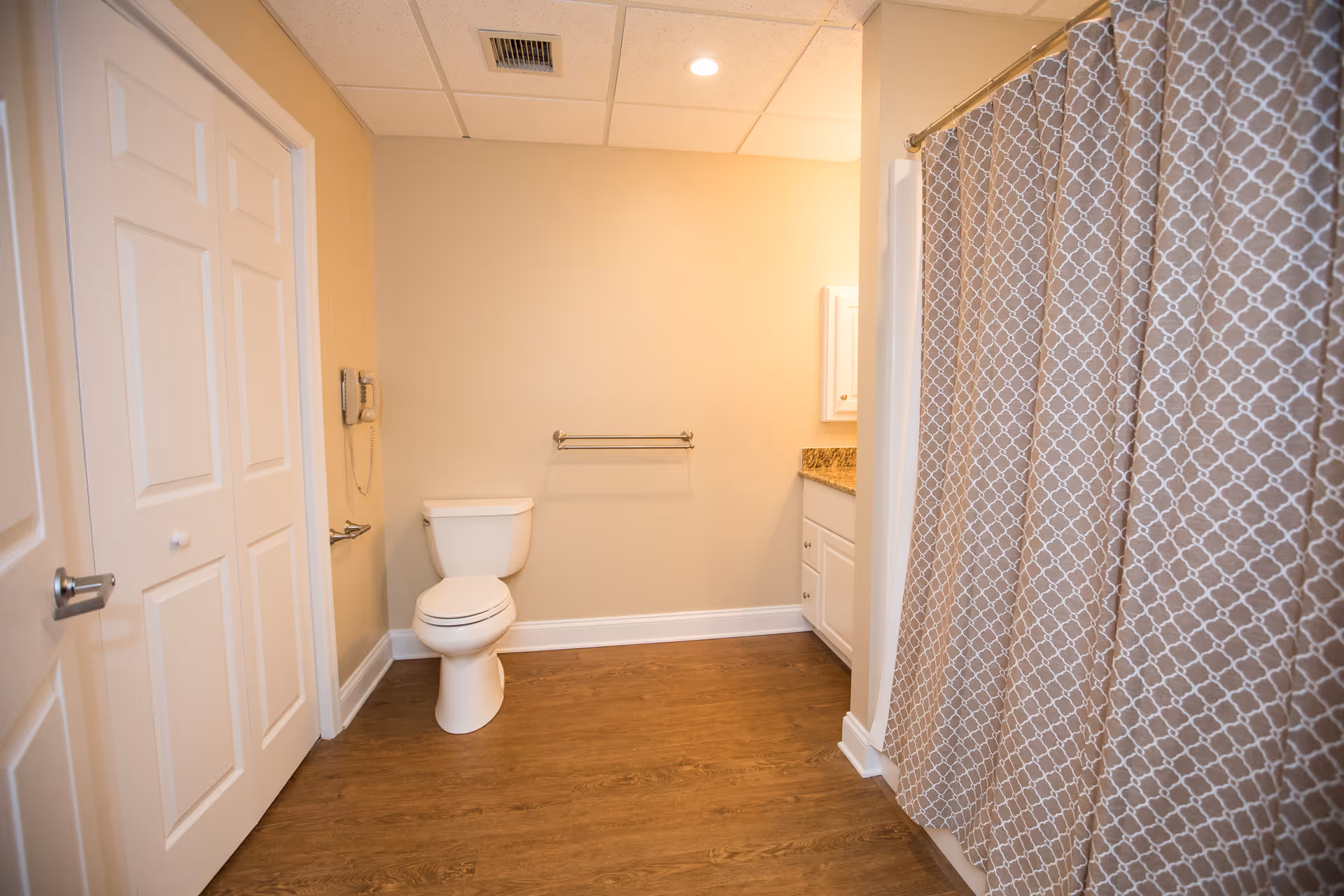A clean bathroom with a white toilet, a towel rack on the wall, a shower with a patterned curtain, a white cabinet with a granite countertop, and wooden flooring.