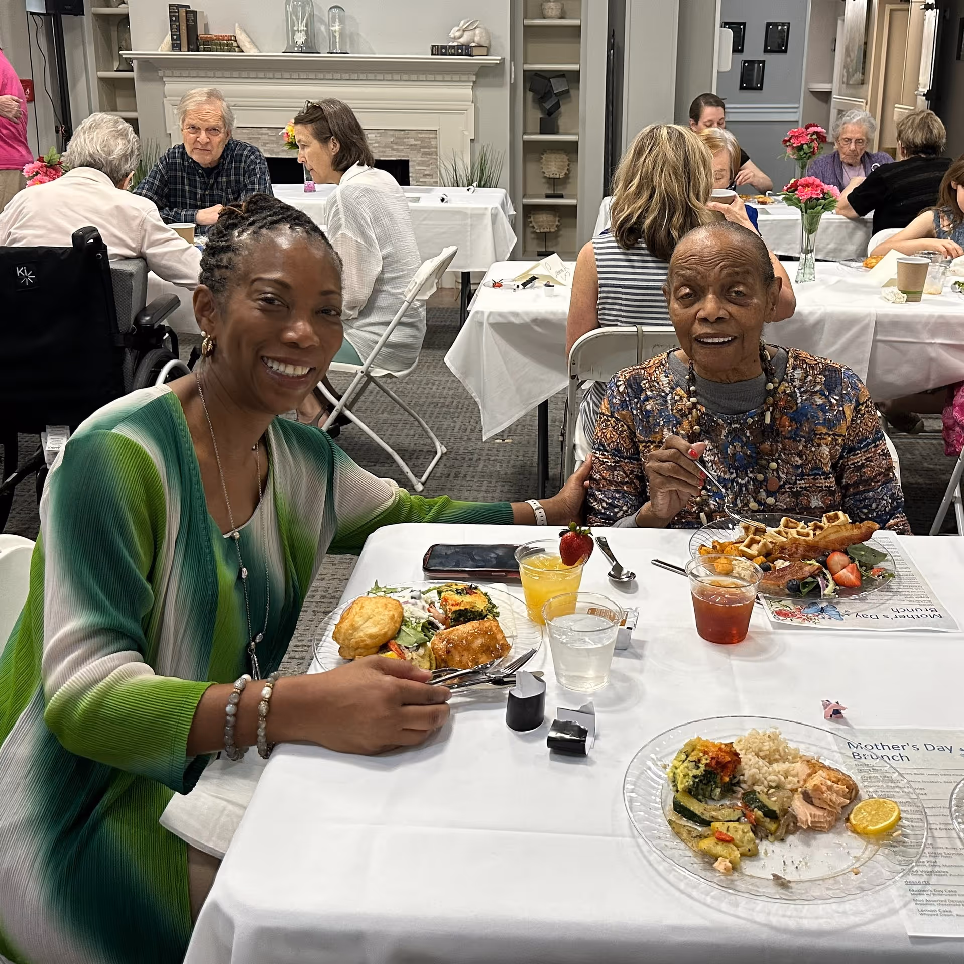 Two women sitting at a table enjoying a meal together in a dining area. The woman on the left is smiling and wearing a green and white top, while the woman on the right is holding a fork and wearing a patterned top. Other people are seated at tables in the background, and the room has a fireplace and shelves with decorative items.