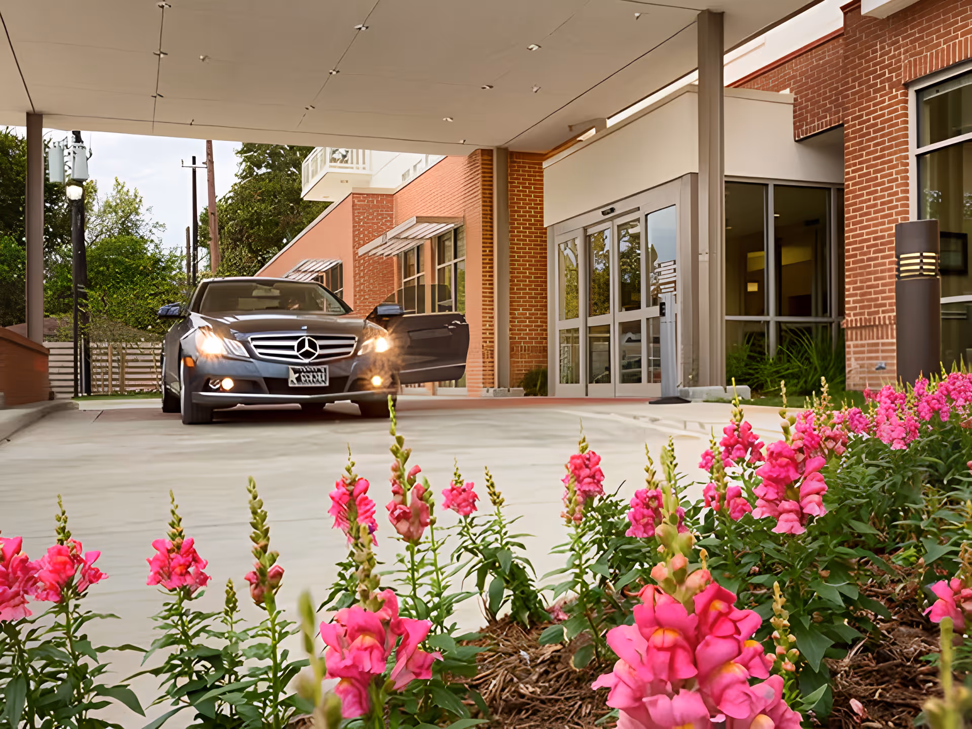 A black Mercedes car with its front door open is parked under a covered entrance of a brick building. In the foreground, there are vibrant pink flowers planted along the driveway leading to the building's glass entrance doors.