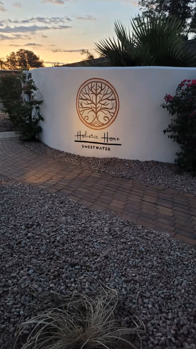 A curved white wall with a circular tree logo and the text 'Holistic Home SWEETWATER' illuminated by a light. The wall is surrounded by gravel and a brick pathway, with some plants and trees visible around it during sunset.
