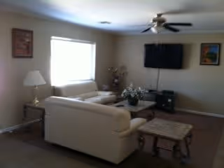 A living room with beige walls and carpet, featuring a white sofa and loveseat arranged around a wooden coffee table with a floral centerpiece. A small side table with a lamp is next to the sofa. A flat-screen TV is mounted on the wall above a media console. Ceiling fan with lights is on the ceiling. Two framed pictures hang on the walls.