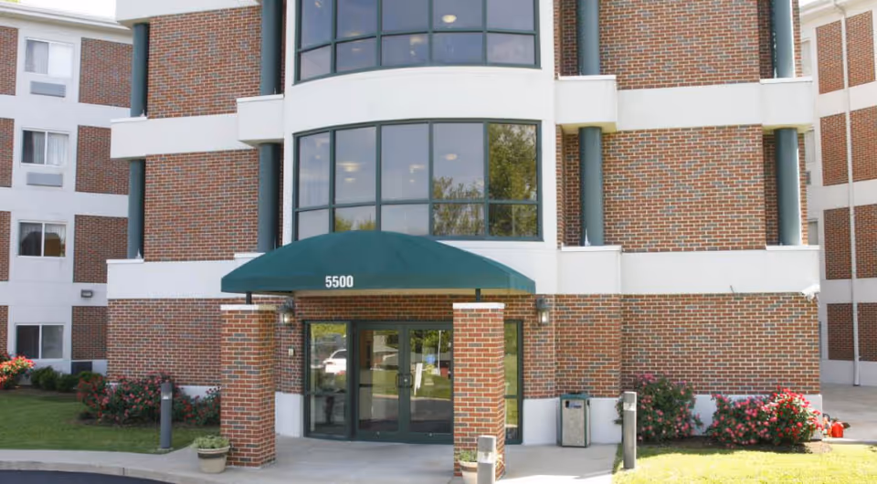 Entrance of a multi-story brick building with large windows and a green awning above the glass double doors. The awning displays the number 5500. There are flower beds with pink flowers on either side of the entrance and a small potted plant near the sidewalk.