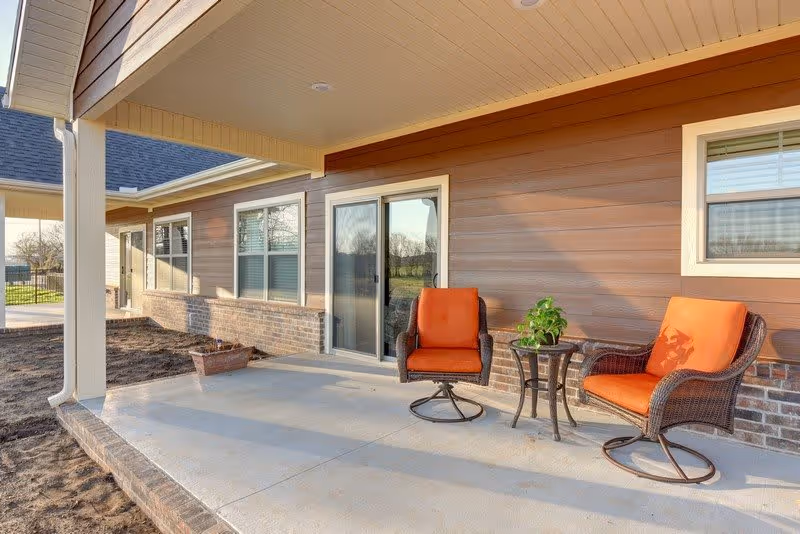 Covered outdoor patio area with two wicker chairs featuring orange cushions and a small round table with a potted plant, attached to a building with brown siding and brick accents, and sliding glass door leading inside.