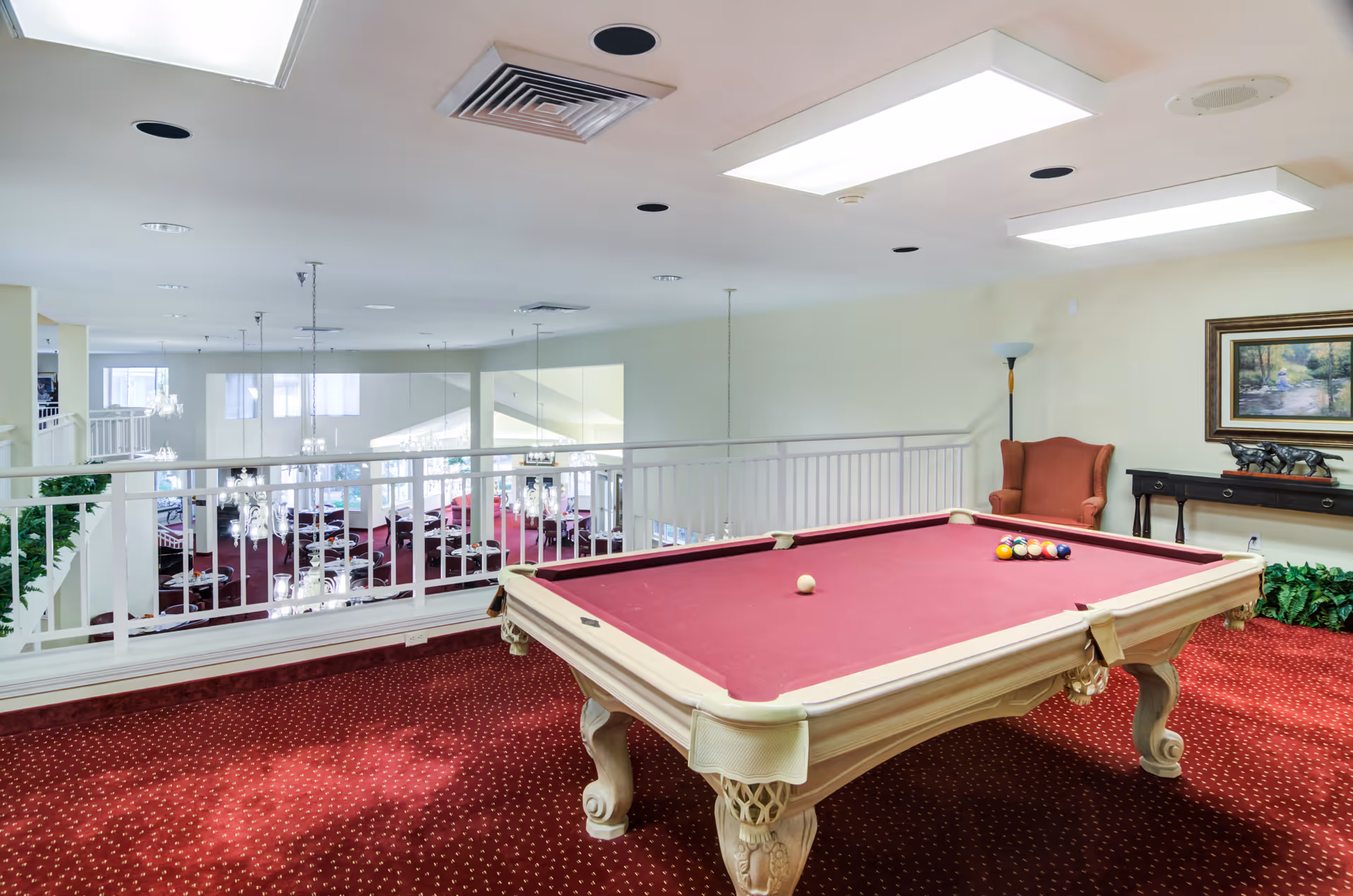 Interior view of a recreational area in Somerset Assisted Living featuring a pool table with a red felt surface and billiard balls arranged on it. The room has a red carpet with a dotted pattern, a wingback chair, a floor lamp, a console table with decorative items, and a framed painting on the wall. The area overlooks a dining room below with multiple tables and chandeliers.