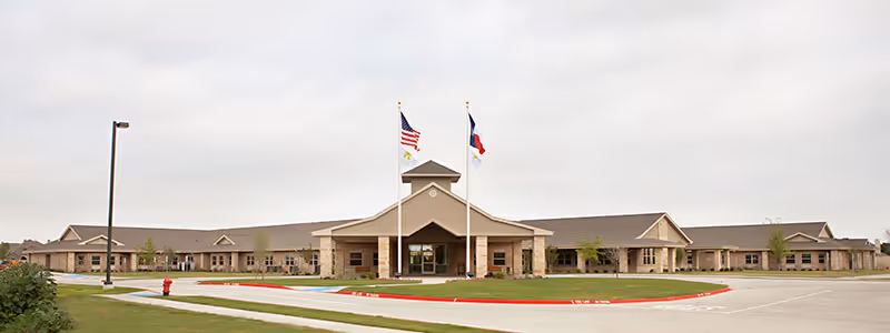 Wide exterior view of a single-story senior living facility building with a covered entrance, two flagpoles displaying the American and Texas flags, a circular driveway, and a well-maintained lawn under a cloudy sky.