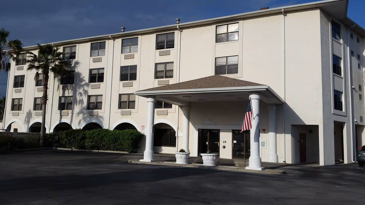 White multi-story senior living building with arched entrances, columns, flag, and palm trees in parking lot.