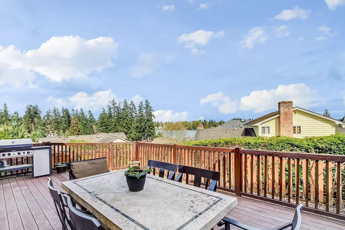 Outdoor wooden deck with a stone table surrounded by chairs, a potted plant on the table, a barbecue grill, and a small refrigerator. The deck overlooks neighboring houses, trees, and a body of water under a partly cloudy sky.