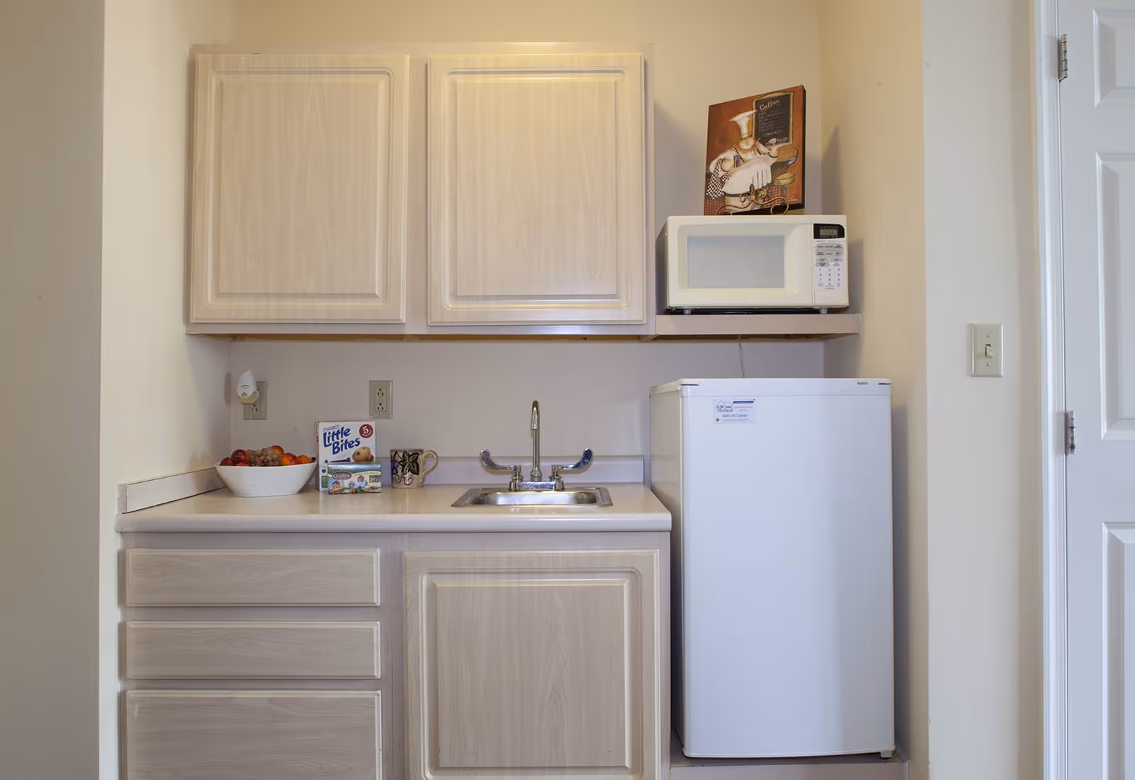 A small kitchenette area with light wood cabinets, a countertop with a sink, a bowl of fruit, a box of Little Bites snacks, and a mug. There is a white mini refrigerator and a microwave on a shelf above it. A decorative sign with a chef illustration is placed on top of the microwave.