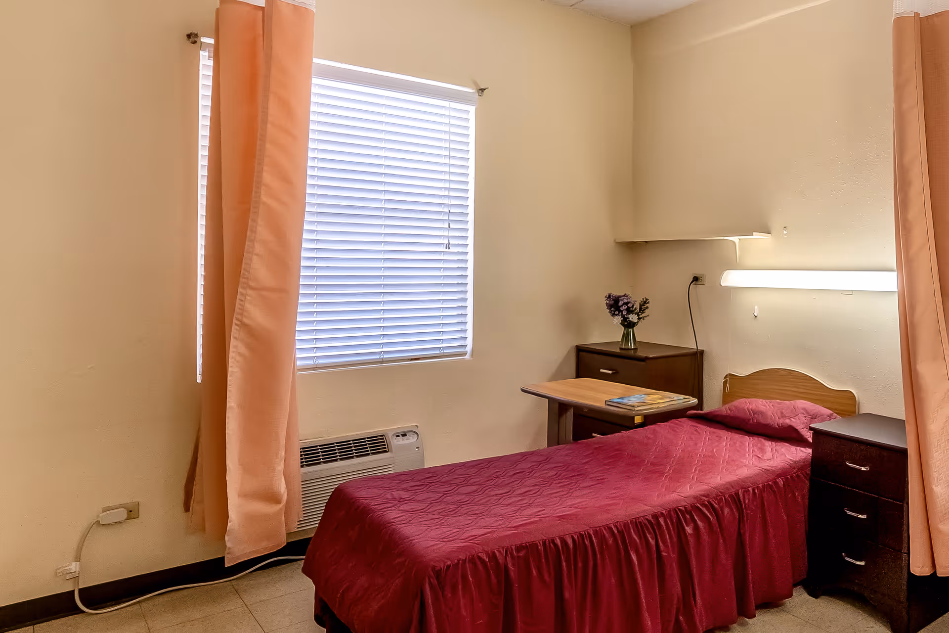 Small nursing facility bedroom with a single bed covered in a maroon bedspread, bedside tables, overbed table, window with blinds and peach curtains.