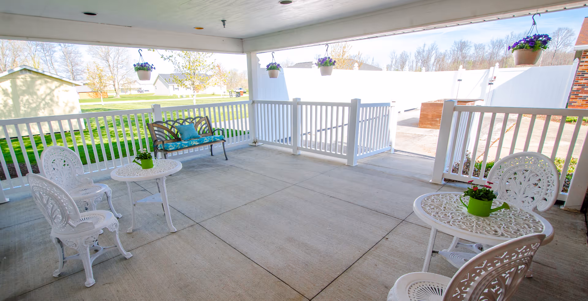 Covered outdoor patio with white metal tables and chairs, a cushioned bench, hanging flower pots, and white railing overlooking a grassy yard.