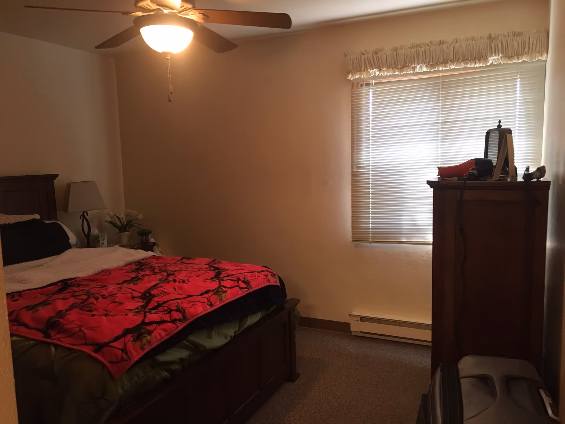A bedroom with a bed, dresser, lamp, blinds, and a ceiling fan.