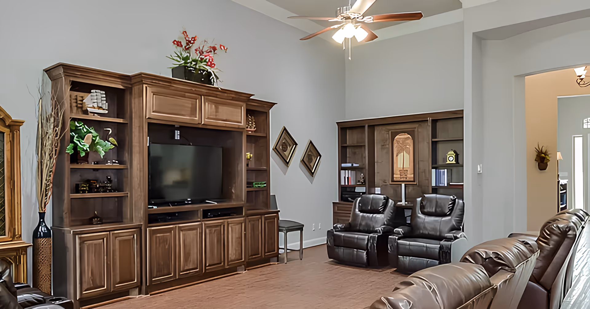 A spacious living room with a large wooden entertainment center housing a flat-screen TV and decorative items. The room features three dark leather recliners, a ceiling fan with lights, and a wooden desk with shelves in the corner. The walls are painted light gray, and there are two diamond-shaped mirrors hanging on one wall.