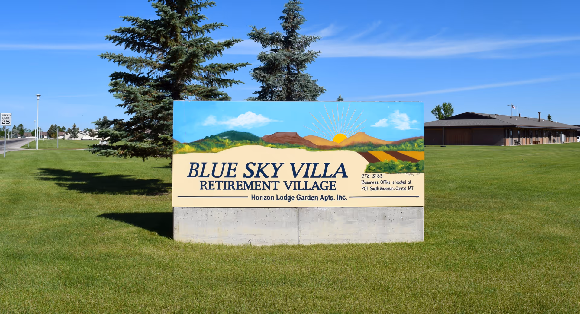 A large sign for Blue Sky Villa Retirement Village with a painted landscape of mountains and a sunrise. The sign is set on a concrete base on a grassy lawn with trees and a building in the background under a clear blue sky.