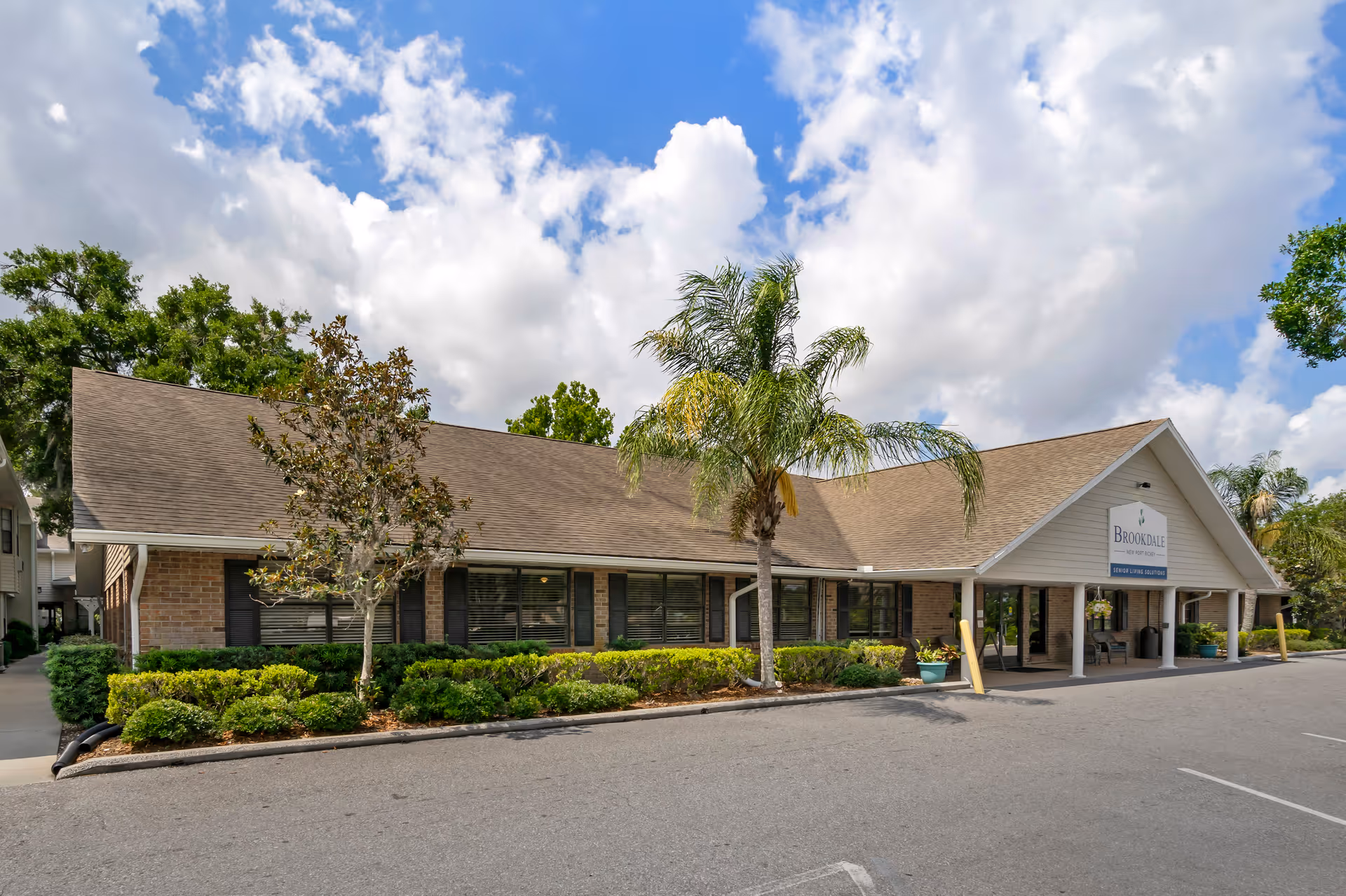 Exterior view of Brookdale New Port Richey senior living facility showing a single-story building with a sloped roof, large windows, and a covered entrance. There are palm trees and shrubs in front of the building under a partly cloudy blue sky.