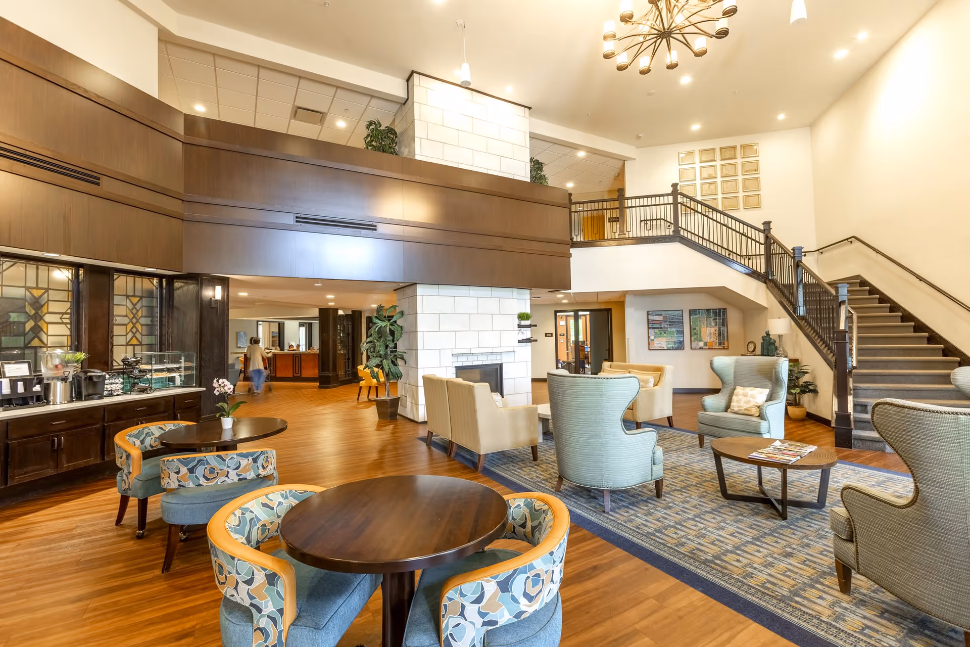 A spacious and well-lit senior living facility common area with wooden floors, comfortable upholstered chairs, round wooden tables, a large rug, a fireplace with a white stone surround, and a staircase leading to an upper level. There are plants and artwork decorating the space, and a person walking in the background near a reception desk.