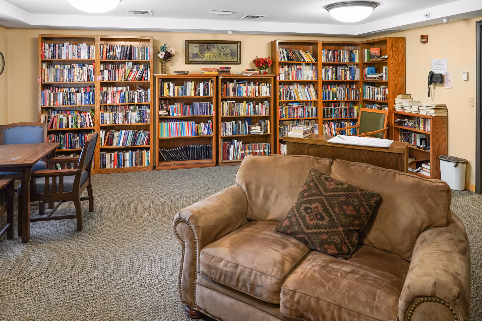 Cozy interior common room with bookshelves lining the back wall, a couch in the foreground, and tables and chairs for reading.