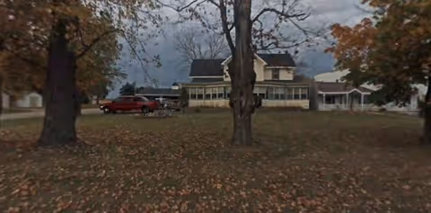 View of a large two-story house with a wraparound porch, surrounded by trees with autumn leaves on the ground under a cloudy sky.