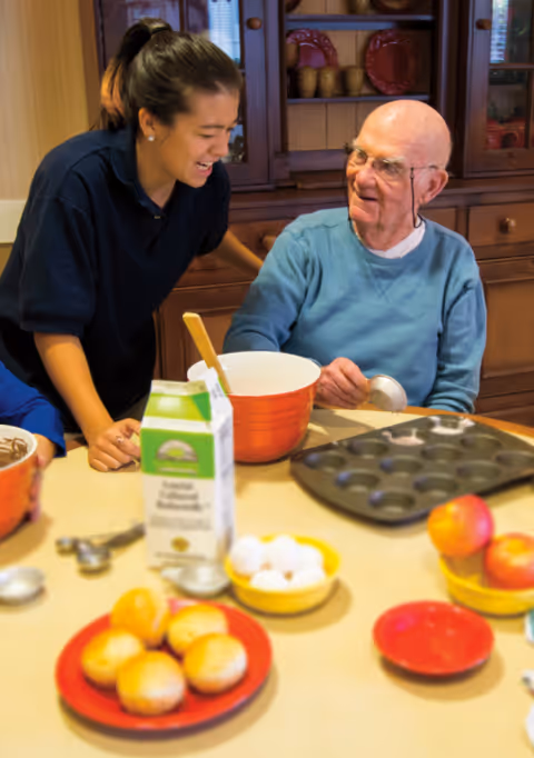 A caregiver and an elderly man smiling while preparing batter at a kitchen table with bowls, a muffin tin, eggs, and fruit.