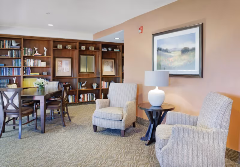 A cozy interior lounge area featuring two upholstered armchairs with a small round wooden table and a white lamp between them. Behind the chairs is a framed landscape painting on a peach-colored wall. To the left, there is a wooden dining table with chairs and a large built-in bookshelf filled with books and decorative items.