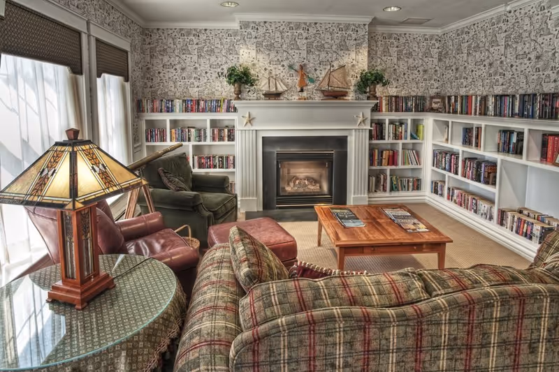Cozy living room with a plaid sofa, green armchair, and red leather chair arranged around a wooden coffee table. The room features a fireplace with decorative sailboats and plants on the mantel, built-in bookshelves filled with books, patterned wallpaper, and a stained glass table lamp on a round table near large windows with blinds.