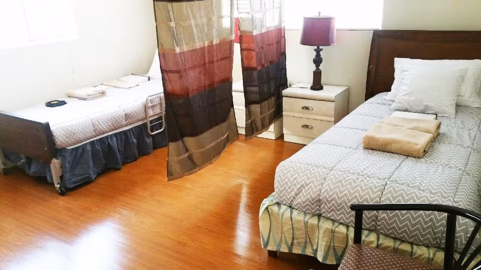 A shared bedroom with two single beds separated by a curtain. Each bed has pillows and folded towels on top. There is a nightstand with a lamp between the beds, and a chair is partially visible in the foreground. The room has wooden flooring and windows letting in natural light.