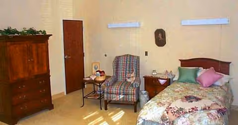 A cozy bedroom with a single bed covered in floral bedding and colorful pillows. Next to the bed is a wooden nightstand with a lamp and some decorative items. There is a plaid upholstered armchair beside a small table holding a few objects. A wooden dresser is positioned against the opposite wall, and a closed door is visible in the background. The room has light-colored walls and flooring.