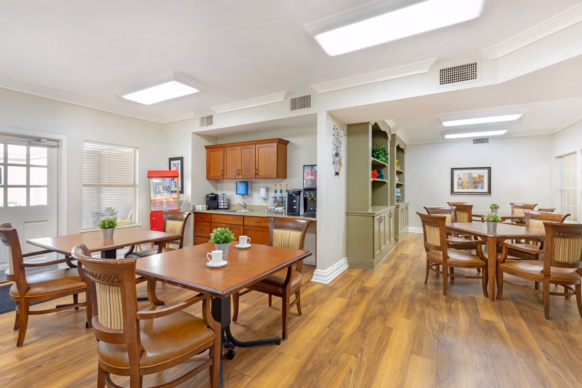 A bright and clean dining area in a senior living facility with wooden tables and chairs. The room features a small kitchenette with cabinets, a coffee machine, and a popcorn machine. There are potted plants on the tables and framed artwork on the walls. The floor is wood, and the ceiling has recessed lighting.