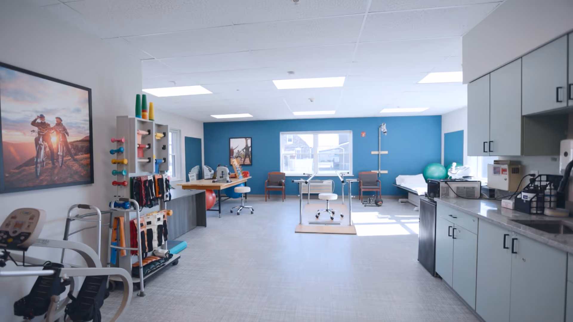 A bright and spacious rehabilitation room with exercise equipment including weights, resistance bands, and therapy balls. There are chairs, a walker, and a treatment table near a large window letting in natural light. The walls are painted white and blue, and there is a large framed picture of two people riding bicycles on the left wall. Cabinets and a countertop with various medical supplies are on the right side.