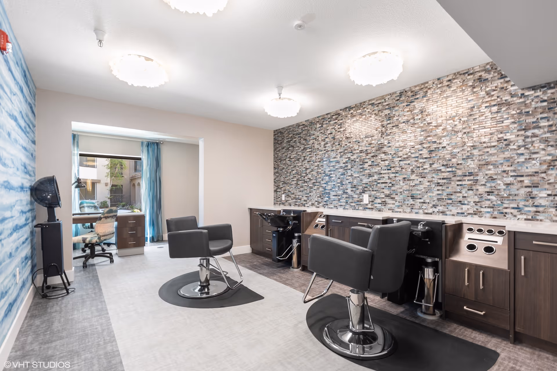 Interior view of a modern hair salon area in a senior living facility with two black salon chairs in front of a mosaic tiled wall with built-in cabinetry and sinks. There is a blue and white patterned wall on the left and a small office area with a desk and chair near a window with blue curtains in the background.