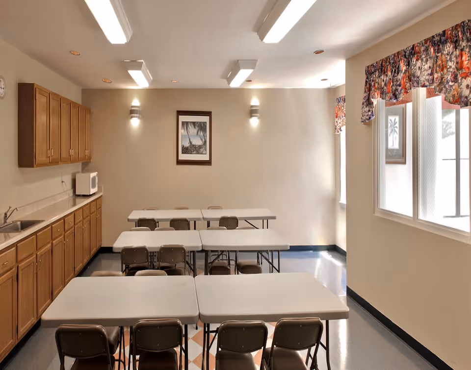 Community dining/activity room with folding tables and chairs, a kitchenette with wooden cabinets and a microwave, framed wall art, and windows with floral valances.