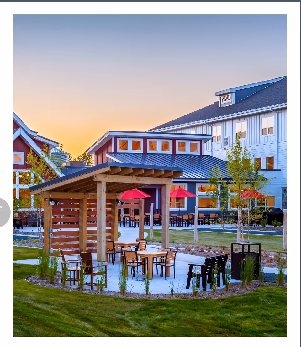 Outdoor seating area at a senior living facility with wooden pergola, tables and chairs, red umbrellas, and well-maintained green lawn with trees and shrubs, set against the backdrop of a multi-story building during sunset.
