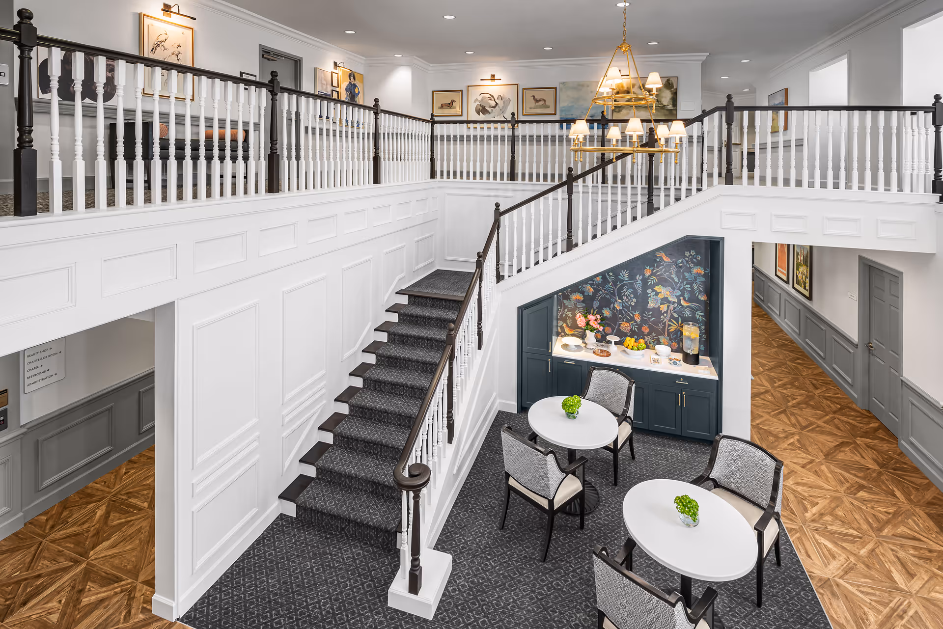 Interior view of a senior living facility featuring a staircase with white and dark wood railings leading to an upper level. Below the staircase is a small seating area with two round tables, each surrounded by four chairs. A decorative cabinet with a floral wallpaper backdrop holds refreshments and snacks. The space is well-lit with wall sconces and a chandelier, and the flooring combines patterned carpet and wood.